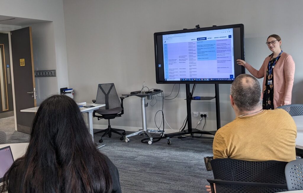 Gillian WIlson stands next to a television screen, which is on a tall stand. Adults are sitting around, watching her presentation. The screen features the SCQF diagram