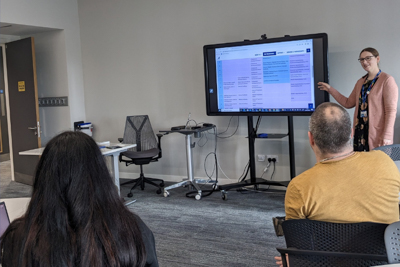 Gillian WIlson stands next to a television screen, which is on a tall stand. Adults are sitting around, watching her presentation. The screen features the SCQF diagram