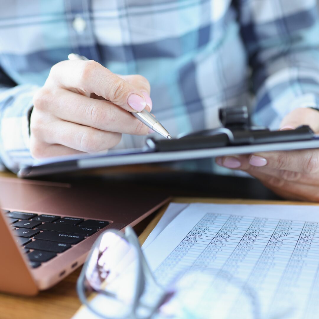 A person in a checked shirt is sitting at a desk, which has a bronze coloured laptop. There's papers on the desk and a pair of glasses. The person is holding a tablet in one hand and a stylus in the other hand, which is pointed towards the tablet.