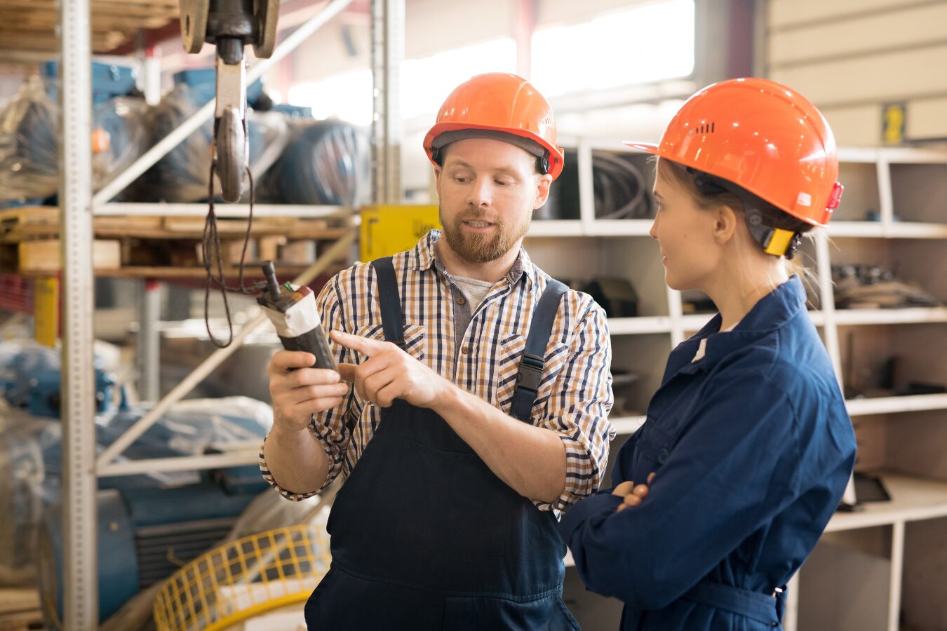A man and woman, both wearing overalls and orange hard hats, are inside a factory. The man is pointing at a hand-held electronic device and talking to a woman. The image signifies the range of qualifications and learning programmes on the SCQF Register