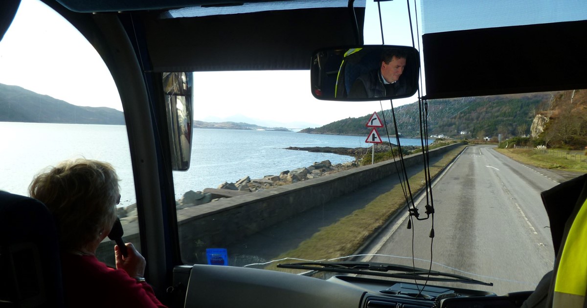 Tourist guide with microphone at front of coach with view of the sea and hills through the windscreen
