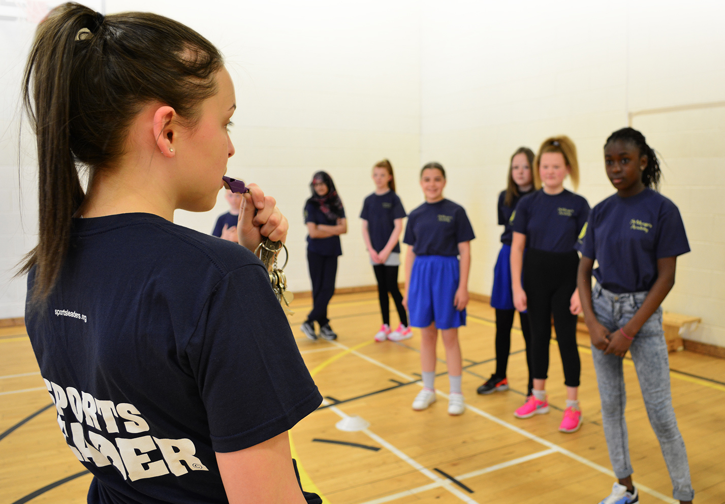 Female Young Leader in gym hall blowing whistle at female pupils