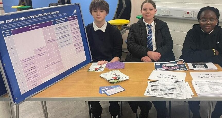 3 Kirkintilloch High School pupils, who are SCQF School Ambassadors, sit at a desk, promoting the SCQF, which shows where the awards, including NPAs, sit