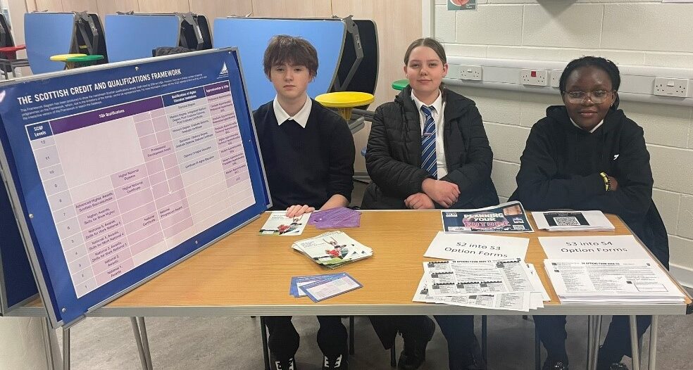 3 Kirkintilloch High School pupils, who are SCQF School Ambassadors, sit at a desk, promoting the SCQF