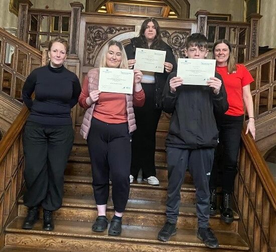 3 Prep for Next Steps learners stand holding their certificates up on a wooden stairway with 2 course tutors.