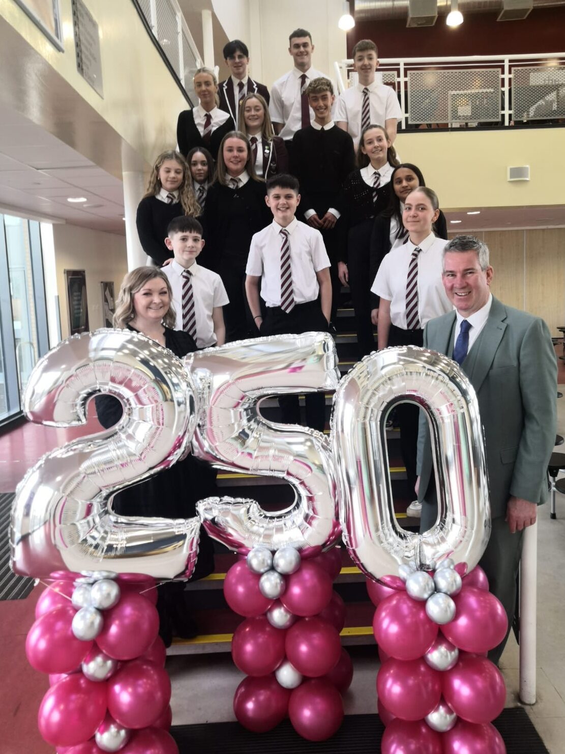 A group of students and staff from Vale of Leven Academy pose for a photo on a staircase inside a school building. The students wear uniforms consisting of white shirts, striped ties, and black or dark-coloured jumpers or blazers. In front of the group, two adults stand beside large silver balloon numbers forming "250," which are supported by pink and silver balloon columns. They are celebrating becoming the 250th SCQF School Ambassador