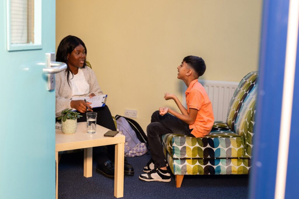 A young boy sits at a table with a young female therapist in a community centre. The boy is engaged in a supportive counselling session, actively participating in the discussion. The therapist is warm and approachable, creating a nurturing environment. The room is brightly lit and welcoming, with comfortable seating, reflecting a safe space for sharing and healing.