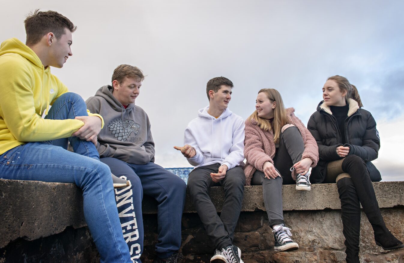 Group of 5 young people sitting on a stone wall outside, chatting, representing the demographic that the SCQF level 5 conflict resolution course is aimed at