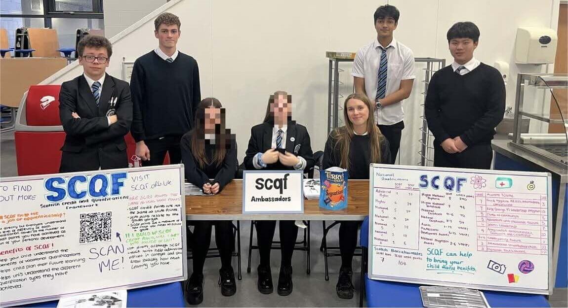 A group of secondary school pupils in school uniform stand and sit behind a table displaying “SCQF Ambassadors” posters and information boards, promoting the Scottish Credit and Qualifications Framework at Douglas Academy.