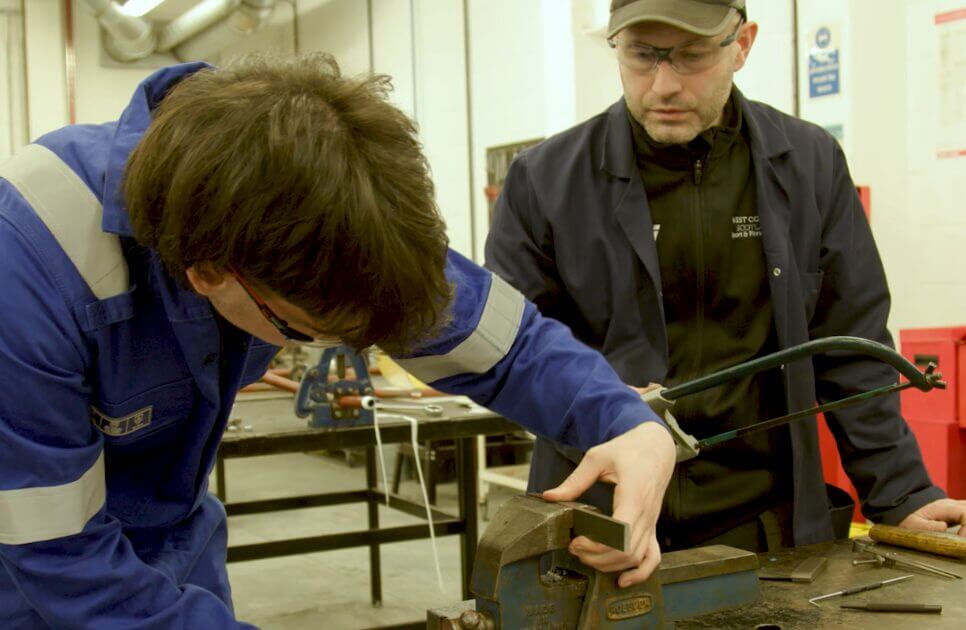 A student in blue protective overalls uses a hacksaw to cut metal secured in a bench vice, while an instructor wearing safety glasses supervises in a workshop training environment at West College Scotland