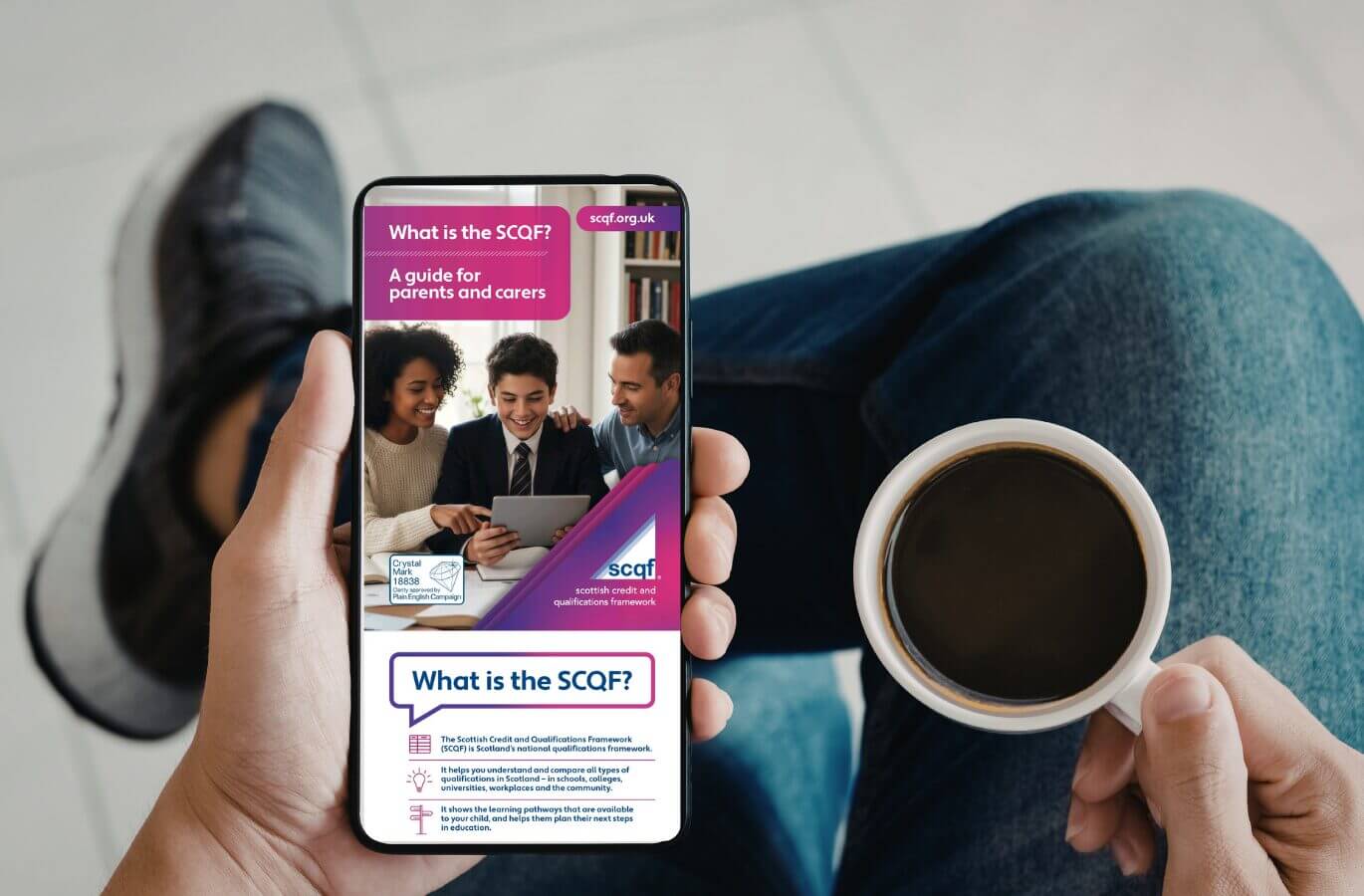 A person holding a smartphone displaying an SCQF leaflet titled “What is the SCQF? A guide for parents and carers.” The leaflet shows a smiling young person with two adults looking at a tablet, alongside SCQF branding and explanatory text. The person is seated, holding a cup of coffee.