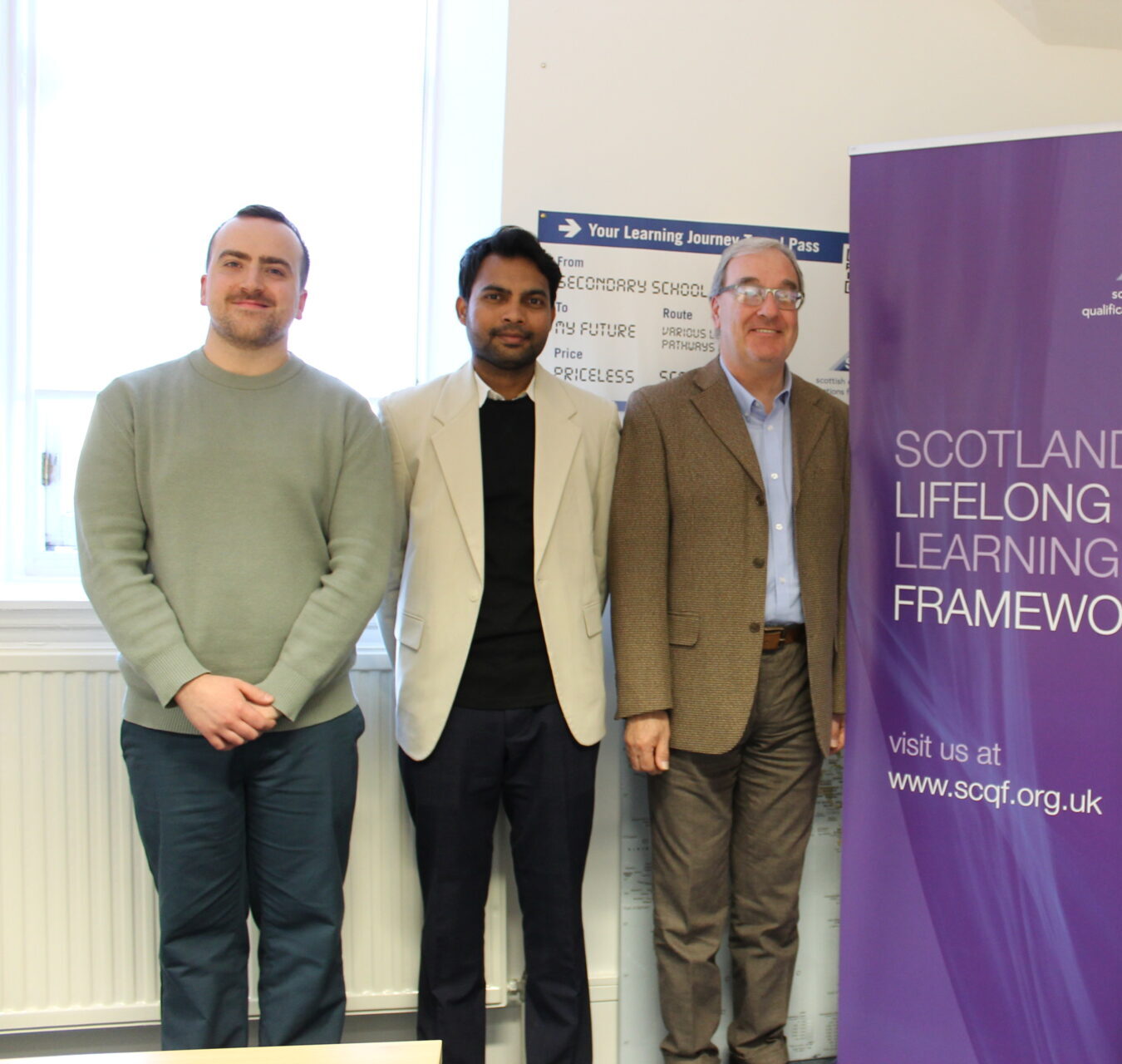3 men stand indoors beside a purple Scottish Credit and Qualifications Framework (SCQF) banner that reads “Scotland’s Lifelong Learning Framework.” They are wearing smart-casual clothing. A window and informational display board are visible in the background.
