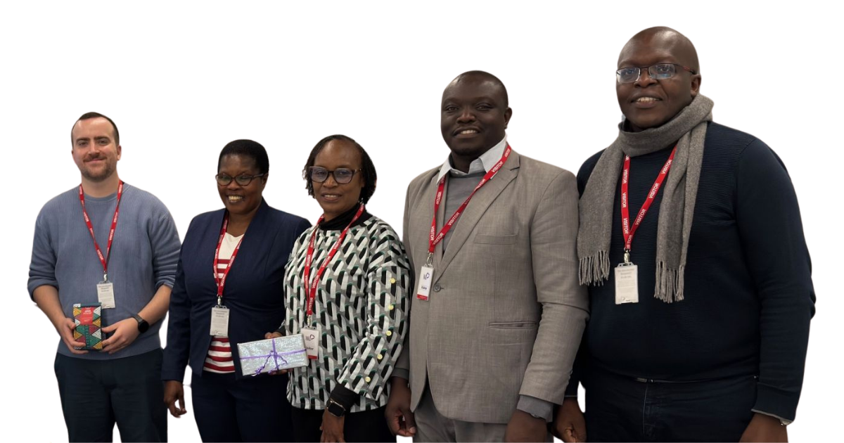 Paul White, SCQFP Lead for International, standing on the left with a group of four visitors from Kenya during a study visit to Forth Valley College. All are wearing conference lanyards and smiling at the camera, with two members of the group holding wrapped gift boxes. The group is dressed in smart-casual to business attire