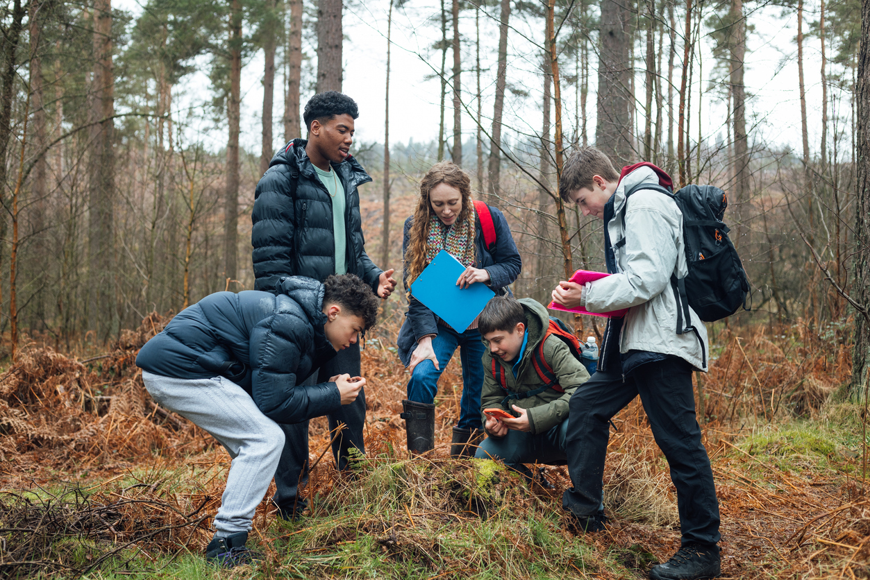 A group of secondary school pupils and a teacher stand together in a woodland area, examining something on the ground as part of an outdoor learning activity. The students wear jackets and backpacks, with some holding clipboards and a tablet, while one kneels to look more closely.