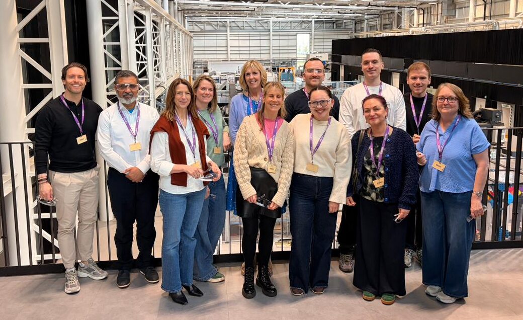 The SCQFP team at NMIS in Paisley stands together on an indoor balcony overlooking a large, modern manufacturing or laboratory space. Around a dozen team members wearing lanyards and name badges face the camera in a single row. The background shows a high-ceilinged industrial environment with white steel beams, exposed piping, and equipment below, creating a bright and structured workspace setting.