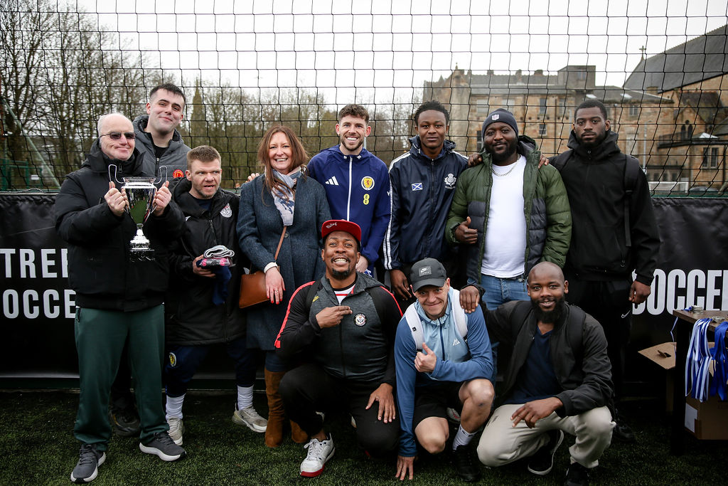 A group of around twelve adults poses together on an outdoor football pitch in front of a netted fence. Several individuals stand in a row at the back while a few kneel or crouch in front. One person near the left side holds a large silver trophy with handles, and another nearby holds a small item. The group is dressed in casual and sporty clothing, including jackets, hoodies, and trainers. One individual in the center wears a blue sports top with the number “8” and a crest. In the foreground, two people crouch, one wearing a cap and pointing toward the camera. To the right side, a small table or stand displays medals with blue ribbons. Behind the fence, trees and a large historic-looking building are visible against an overcast sky.