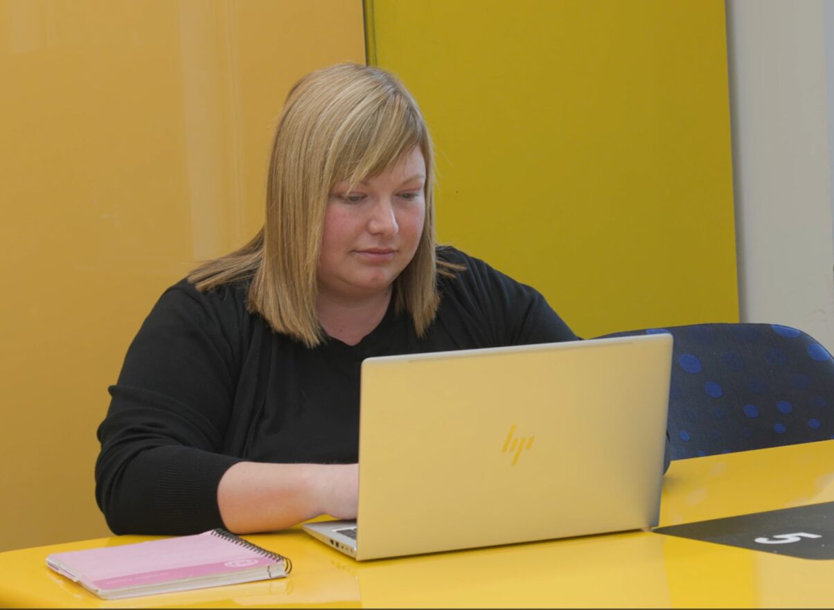 A person sits at a bright yellow table using a silver HP laptop. A pink notebook lies beside the laptop. The background features a yellow wall and a blue chair with a dotted pattern. The image relates to a student, Stacey Grieve, who is studying a Masters in Community Development and Youth Work at the University of Dundee. She features as part of a Recognition of Prior Learning case study.