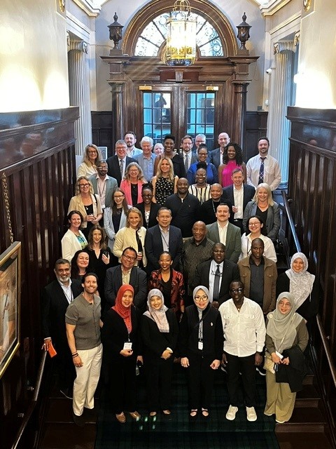 A large group of adults in business attire poses on a wooden staircase inside an ornate building, with a chandelier and arched window behind them.