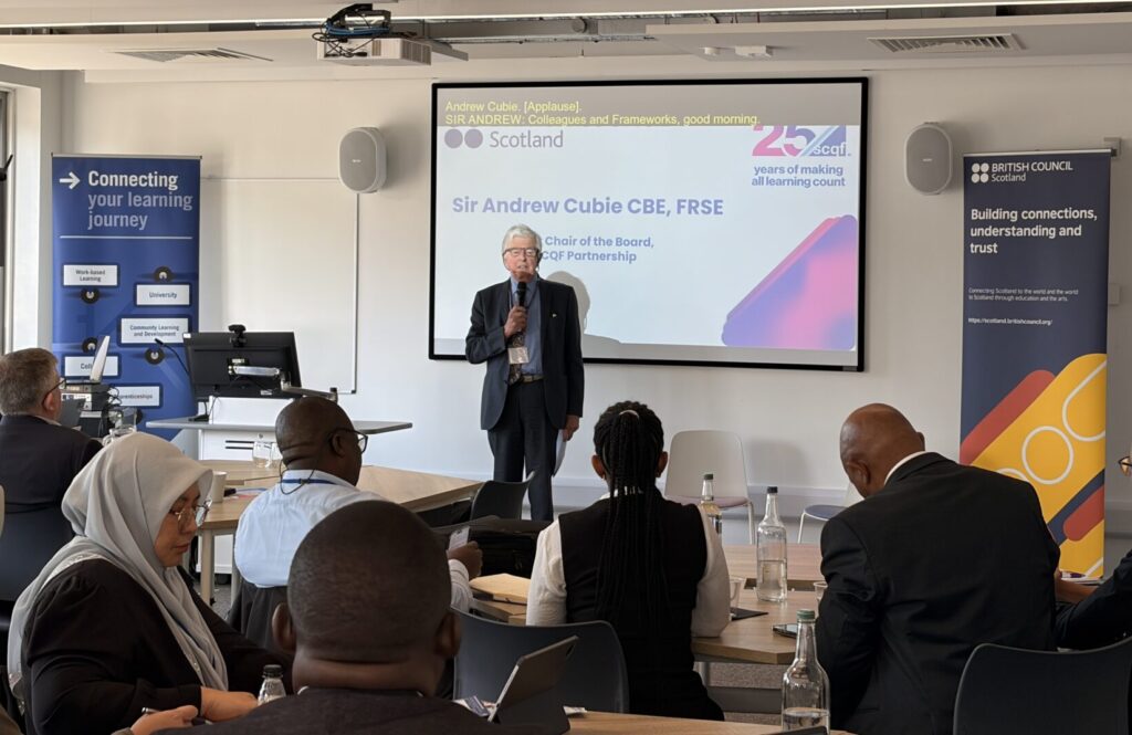 Sir Andrew Cubie stands at the front of a lecture room at Glasgow Caledonian University, addressing an audience seated at tables, with a presentation slide and British Council banners behind them.