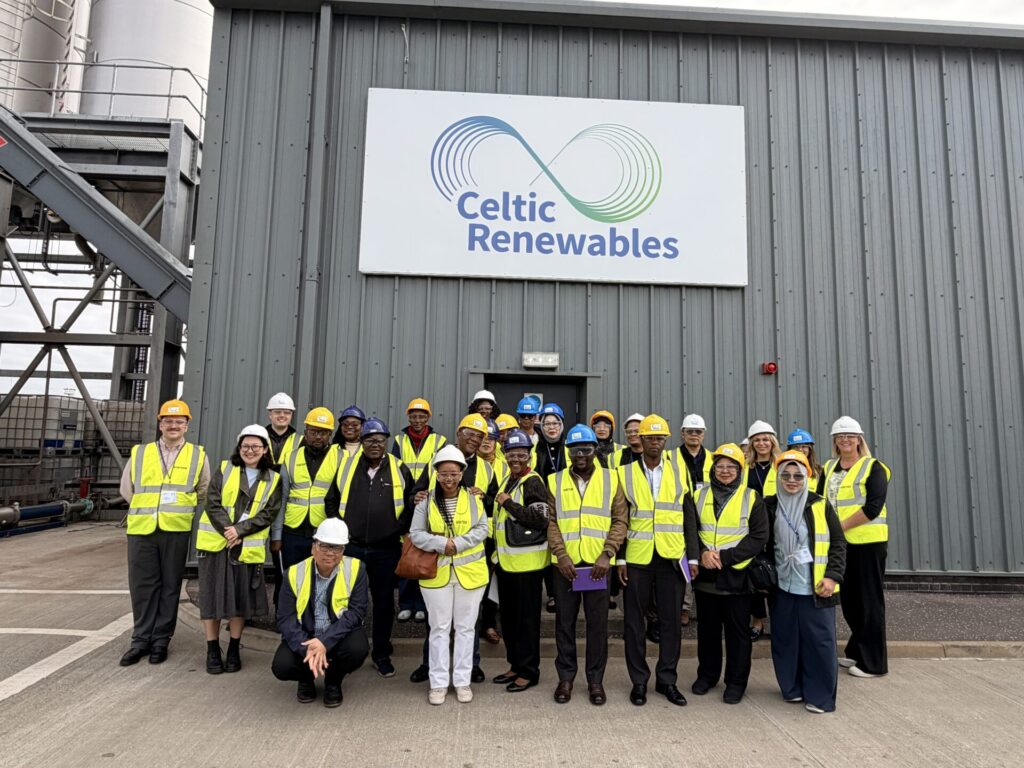 A large group study visit delegates wearing high-visibility vests and safety helmets stands outside an industrial building beneath a sign reading “Celtic Renewables,” posing for a group photo.