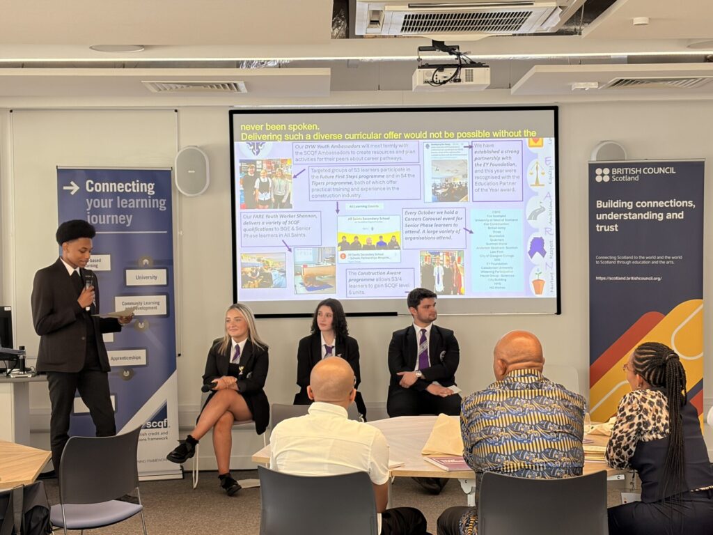 An All Saints Secondary student in a school uniform stands at the front of a lecture room speaking into a microphone, while three other students sit beside them facing an audience, with a presentation slide and British Council banners behind them.