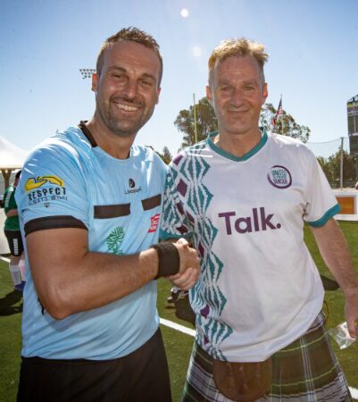 Two people are standing outdoors on a sunny day on what appears to be a sports field with green artificial turf. The two people are positioned in the foreground, facing the camera, and clasping hands in a handshake at the centre of the image.
The person on the left is wearing a light blue athletic shirt with short sleeves and dark trim across the chest and shoulders. The shirt includes multiple logos and patches; one visible patch on the upper sleeve reads “Respect.” The person also wears a dark wristband.
The person on the right, who is Paddy Maloney, is wearing a white sports jersey with green and purple decorative patterns running diagonally across the front. The jersey features the word “Talk.” printed prominently across the chest in bold lettering. The person is also wearing a tartan kilt and has a small sporran (a pouch) hanging at the front. In the right hand, this person is holding a clear plastic water bottle.
In the background, there are elements of a sports venue: a goalpost or field structure, a white tent, and some banners or flags. The sky is clear and bright blue with strong sunlight, creating distinct shadows and highlights on the subjects.