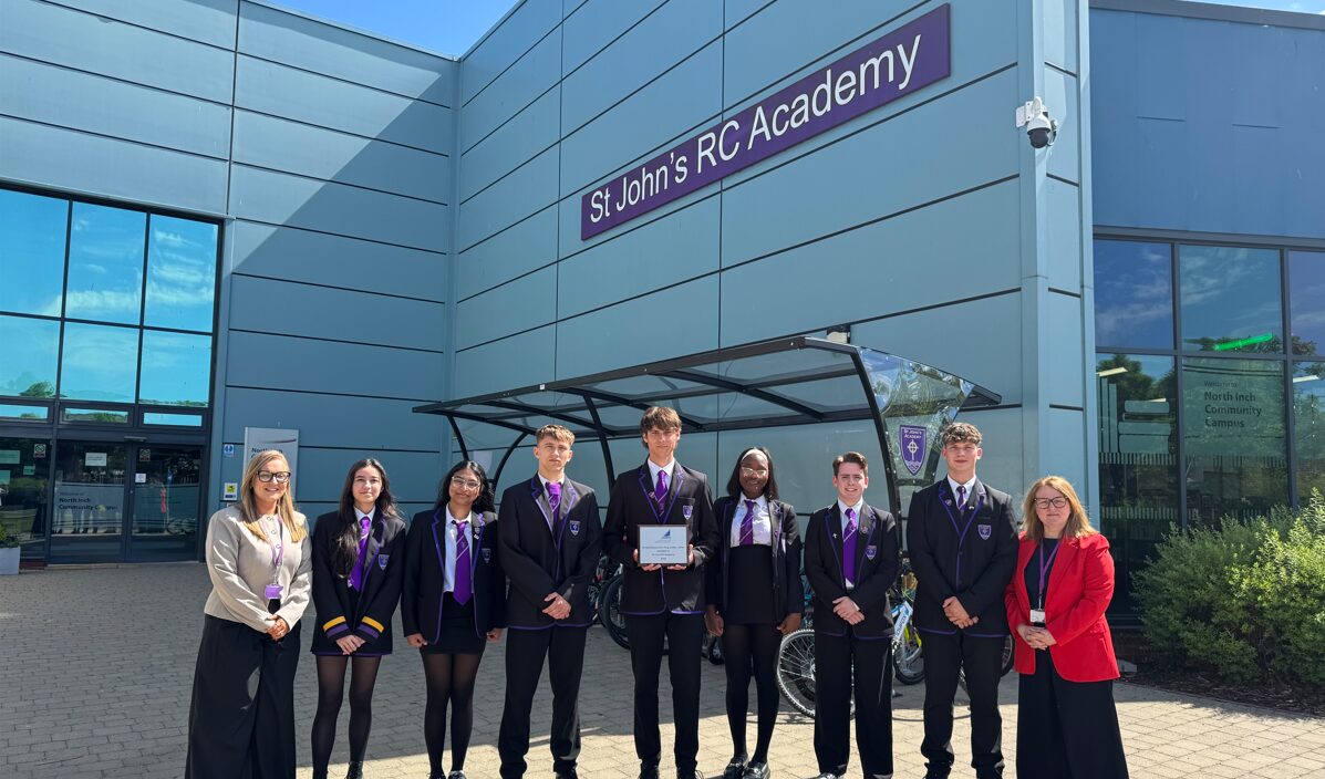 A group of nine individuals stand in a line outdoors in front of a modern grey building with metal panelling. Above them, a large purple sign reads “St John’s RC Academy.” The group is positioned beneath a glass-and-metal bike shelter, with several bicycles visible behind them. Eight individuals in the centre are wearing matching school uniforms consisting of dark blazers with purple trim, white shirts, and purple ties. One individual in the middle holds a rectangular plaque or certificate, suggesting an award or recognition. On either side of the group stand two adults in professional attire—one on the far left wearing a light blazer and dark skirt, and one on the far right wearing a bright red blazer and dark trousers. The building includes large windows reflecting blue sky and clouds. To the right, a sign on a glass panel reads “North Inch Community Campus.” Bushes and greenery are visible near the building, and the ground is paved. The sunlight is strong, casting clear shadows, indicating a bright day.