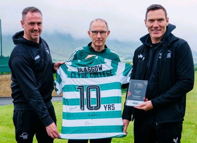 Three people (from left, Glasgow Clyde College curriculum manager David McGurn, Celtic FC manager Martin O'Neill, and Glasgow Clyde College sports lecturer Shaun Gallagher} stand outdoors on a grassy area, holding a green and white striped Celtic FC jersey and a plaque. The jersey displays the text “Glasgow Clyde College” and “10 YRS,” with hills and a cloudy sky in the background.