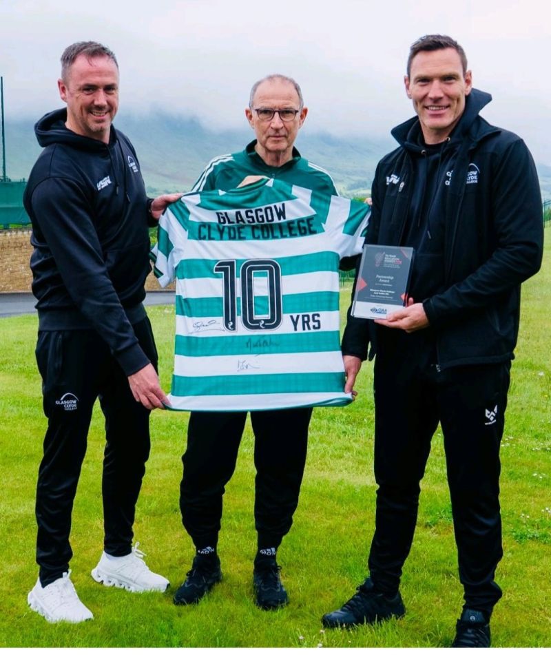 Three people (from left, Glasgow Clyde College sports lecturer Shaun Gallagher, Glasgow Celtic FC manager Martin O'Neill and Glasgow Clyde College curriculum manager David McGurn} stand outdoors on a grassy area, holding a green and white striped Celtic FC jersey and a plaque. The jersey displays the text “Glasgow Clyde College” and “10 YRS,” with hills and a cloudy sky in the background.