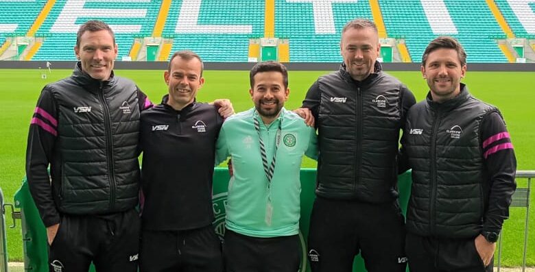 Five people stand together pitchside at Celtic Park stadium in Glasgow. Glasgow Clyde College curriculum manager David McGurn is on the far left and Glasgow Clyde College sports lecturer Shaun Gallagher is second from the right. The group stands behind a metal crowd barrier with the football pitch and the stadium’s green-and-white seating behind them. Large white letters spelling “CELTIC” are visible across the stand, with an Adidas logo above. Two foreground signs display the text “PLEASE MIND.” The photograph is taken from pitch level under the covered stadium roof.