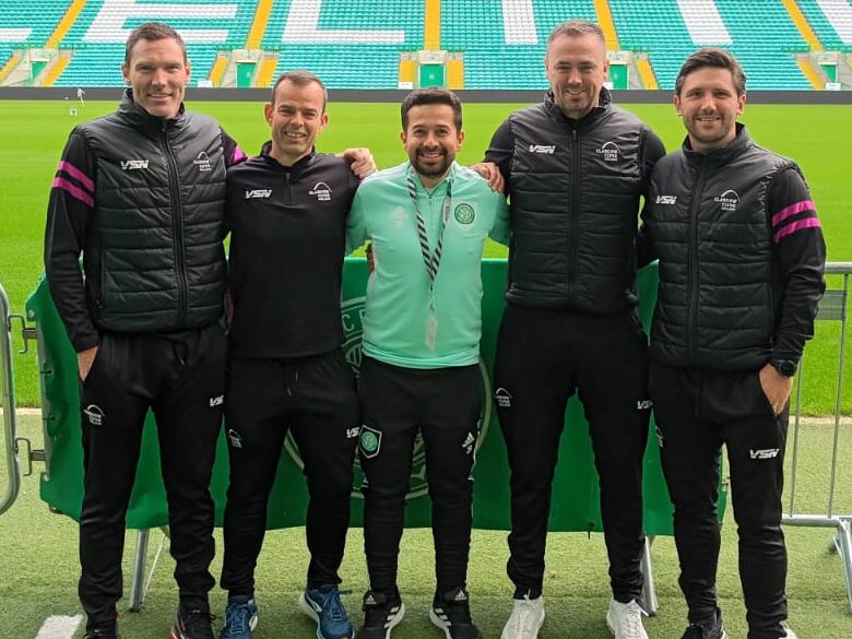 Five people stand together pitchside at Celtic Park stadium in Glasgow. Glasgow Clyde College curriculum manager David McGurn is on the far left and Glasgow Clyde College sports lecturer Shaun Gallagher is second from the right. The group stands behind a metal crowd barrier with the football pitch and the stadium’s green-and-white seating behind them. Large white letters spelling “CELTIC” are visible across the stand, with an Adidas logo above. Two foreground signs display the text “PLEASE MIND.” The photograph is taken from pitch level under the covered stadium roof.