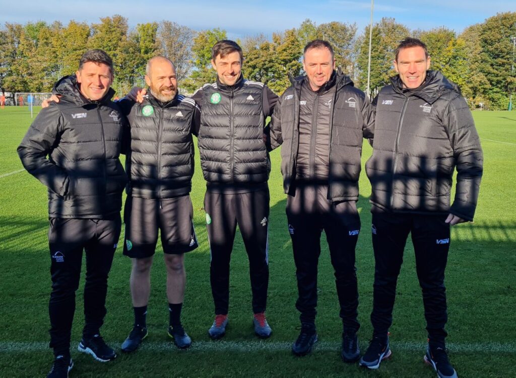 Five people wearing matching black Celtic FC jackets and tracksuit bottoms stand arm in arm on a grass football pitch. Goalposts and trees are visible in the background under a bright blue sky, with sunlight casting long shadows across the field.