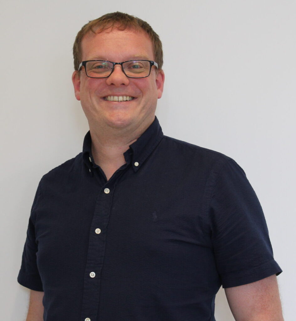 Rob Stroud wearing rectangular glasses and a dark navy short-sleeved button-up shirt stands against a plain white wall. The person is facing the camera, with the upper body visible in a well-lit indoor setting.