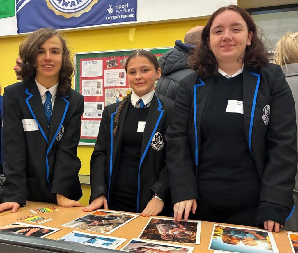 Three Castlemilk High School students in school uniforms stand behind a table displaying photographs and project materials at an SCQF School Ambassador event. A display board of student work is behind them. 