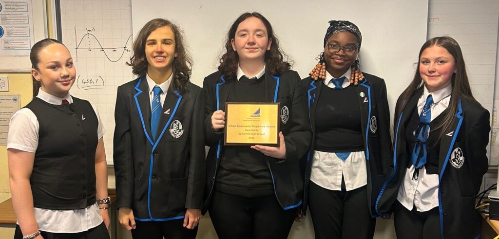 Five Castlemilk High School in school uniforms stand together in a classroom, with one student holding a bronze SCQF School Ambassador Programme award plaque. A whiteboard with a maths graph and classroom displays are visible in the background.