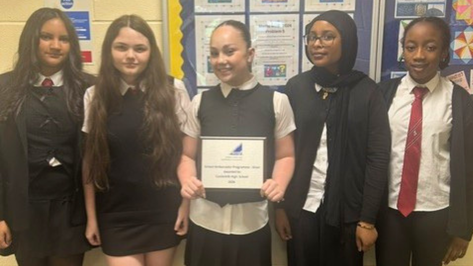 Five Castlemilk High School students stand together in a school corridor, with one student in the centre holding a silver SCQF Ambassador plaque. They are smiling for the camera, with school display boards and examples of students’ work visible behind them.
