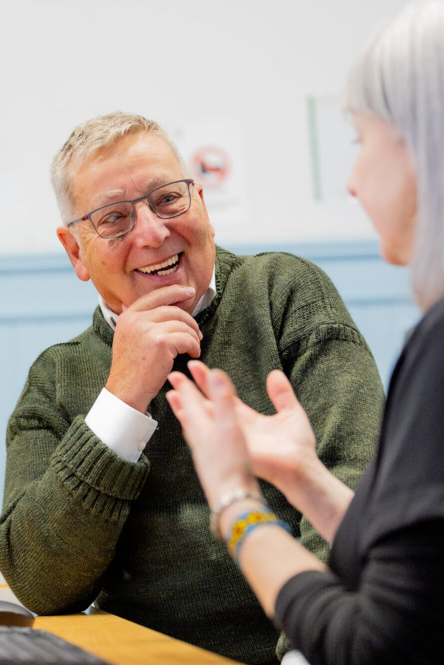 Charles Clark wearing a green jumper and glasses sits at a desk in a learning or office environment, engaged in conversation with another person across the table. Computer keyboards, a mouse and a whiteboard are visible, suggesting a training, mentoring or classroom setting. The image is brightly lit and focuses on the interaction between the two individuals.