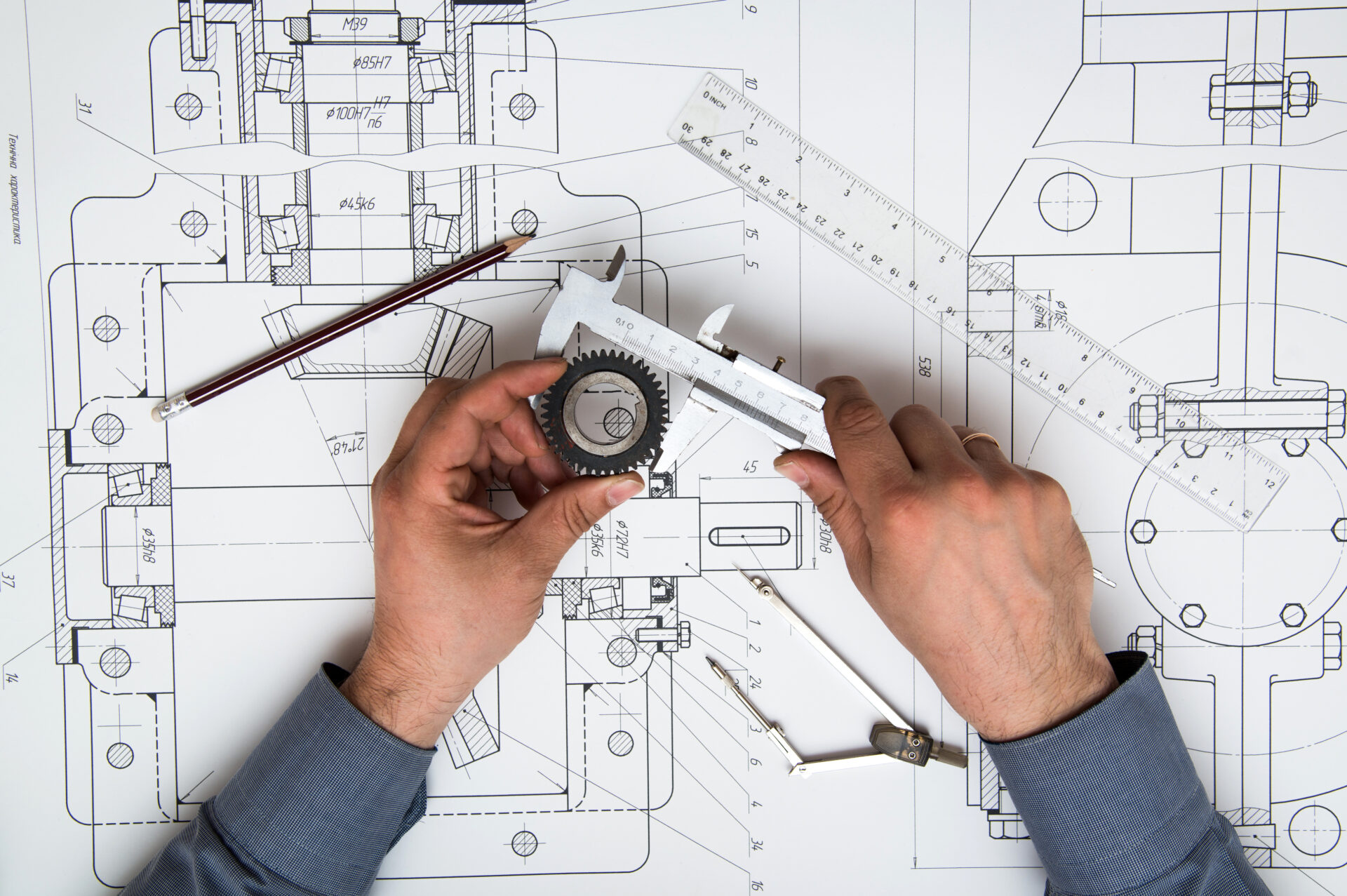 Hands use a caliper to measure a gear placed on a detailed engineering drawing, with technical drafting tools and plans spread across the workspace.