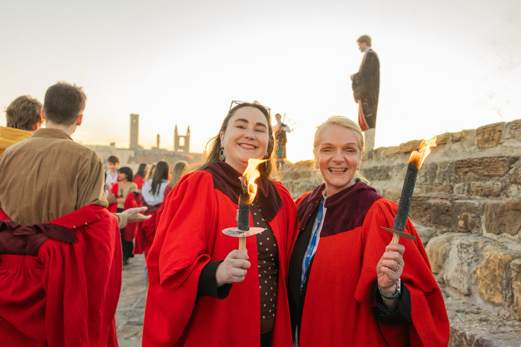 Amanda and Natalie Gaudie wearing bright red academic-style robes stand on a historic stone walkway holding lit ceremonial torches. Several other robed participants and visitors are gathered nearby, while the ruins and skyline of a historic town are visible in the background. The scene is illuminated by warm evening sunlight, creating a celebratory atmosphere during an outdoor academic or ceremonial event.