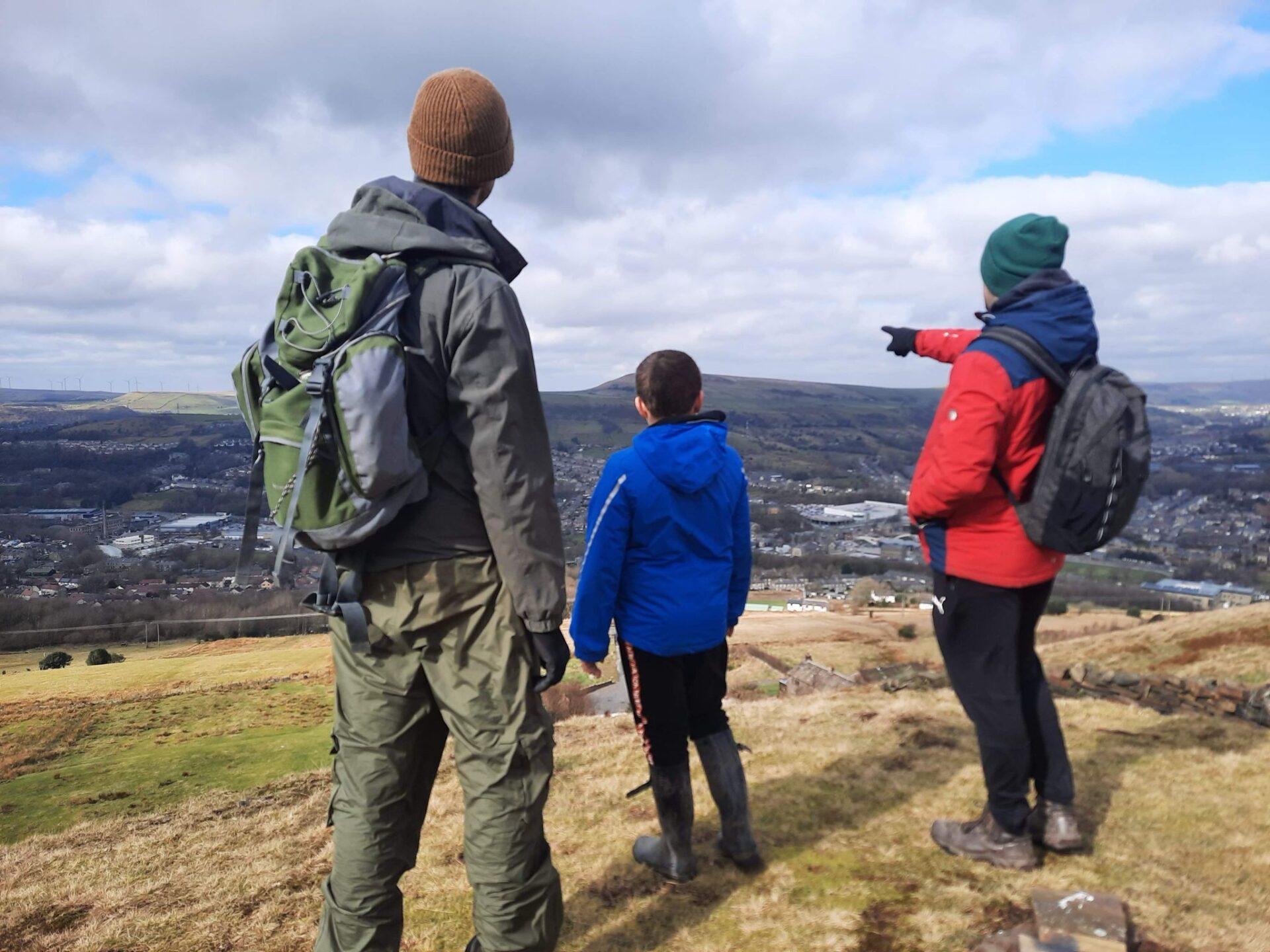 Three people in outdoor clothing stand on a grassy hillside overlooking a valley and distant hills. One person wears a large green backpack, another wears a blue jacket, and a third wears a red jacket and points toward the landscape. The scene is lit by daylight under a partly cloudy sky, with a town, fields, and rolling hills visible below.