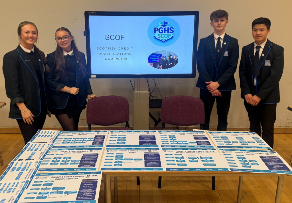 Four Port Glasgow High School students in school uniforms stand around an SCQF information display at a school event. A large digital screen in the centre shows the Scottish Credit and Qualifications Framework (SCQF) logo and presentation materials. Tables in the foreground are covered with subject pathway posters and qualification information charts. The display appears to be promoting awareness of learning pathways, qualifications and the SCQF within a school setting.