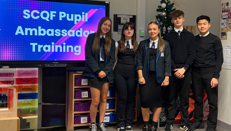 Five Port Glasgow High School pupils stand beside a large digital screen displaying “SCQF Pupil Ambassador Training” in a classroom. The pupils are wearing school uniforms and are positioned in front of storage units and classroom displays. Educational posters are mounted on the wall above the screen, and a decorated Christmas tree stands nearby. The image shows a group participating in training related to becoming SCQF Pupil Ambassadors.