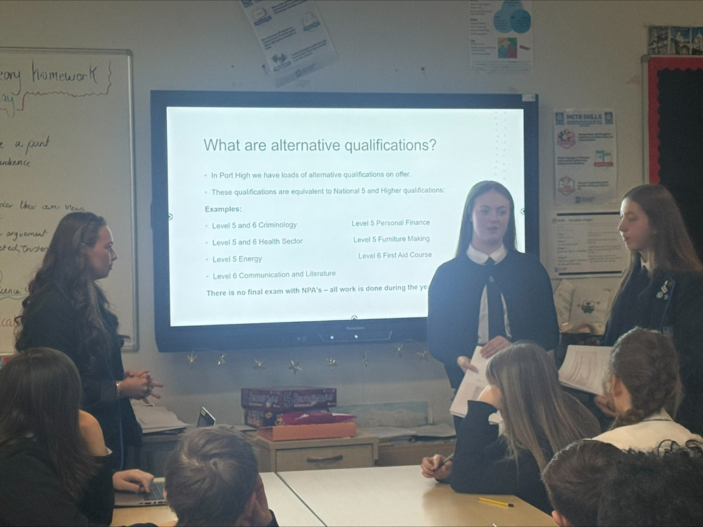 3 Port Glasgow High School students deliver a classroom presentation about alternative qualifications, standing in front of a large digital screen displaying information on qualifications available at Port Glasgow High School. The slide includes examples such as Criminology, Health Sector, Personal Finance, Furniture Making and First Aid courses. Other students are seated around tables listening to the presentation, while classroom whiteboards and educational posters are visible in the background.