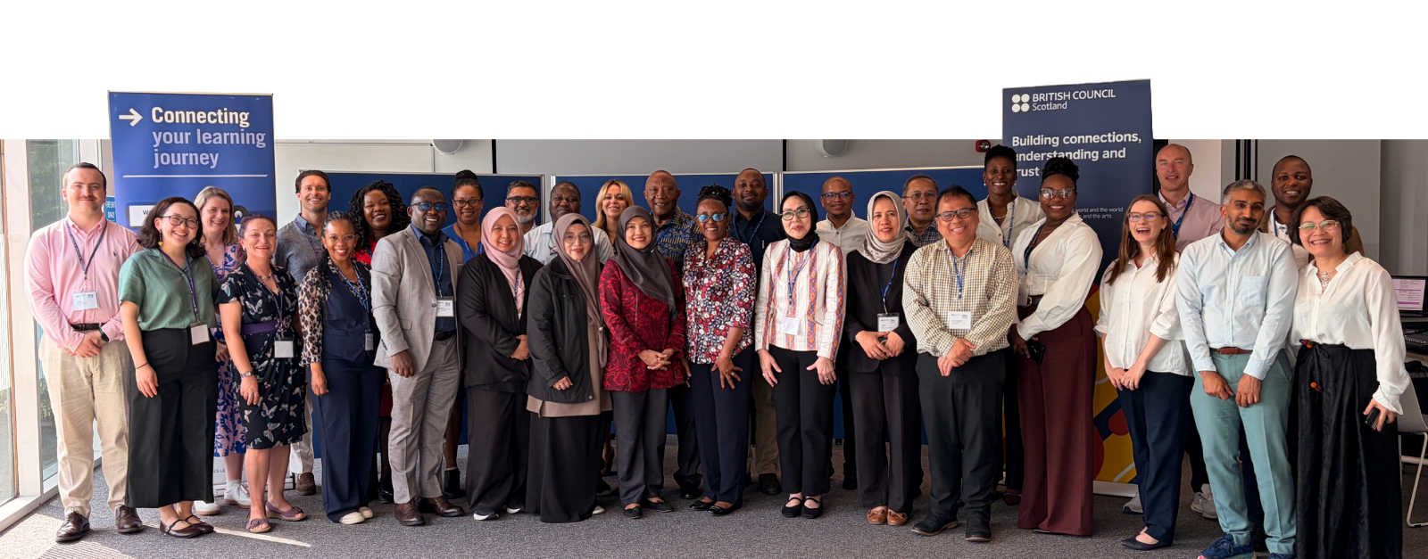 Group photo of approximately 30 delegates and staff standing in a modern meeting room at Glasgow Caledonian University. The group is arranged in several rows in front of a large projection screen, with SCQFP and British Council banners on either side. Large windows on the left let in bright natural light, and attendees wear a mix of business and smart-casual clothing with conference lanyards. Visible banner text includes “Connecting your learning journey” and “Building connections, understanding and trust.”