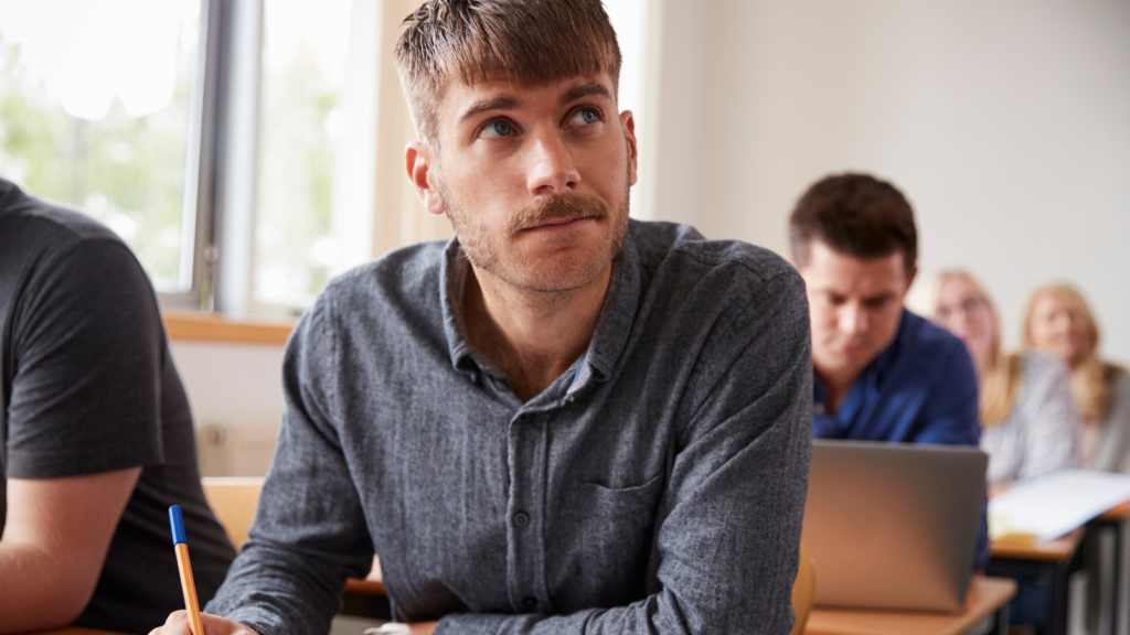 An adult learner sits at a desk in a classroom or training environment, holding a pen and looking towards the front of the room. The learner is wearing a grey button-up shirt and is seated among other learners, some of whom are working on laptops in the background. Large windows provide natural light, creating a bright and welcoming learning setting. The image represents adult education, lifelong learning, skills development, or returning to learning later in life.